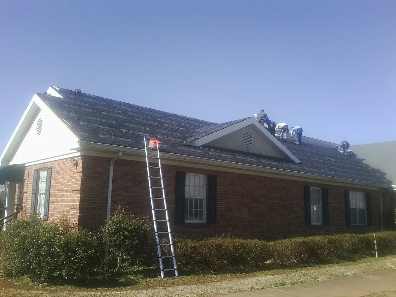 Crew tearing shingles off a brick ranch roof with a ladder set — Emergency Roof Repair work by our crew in Rochester
