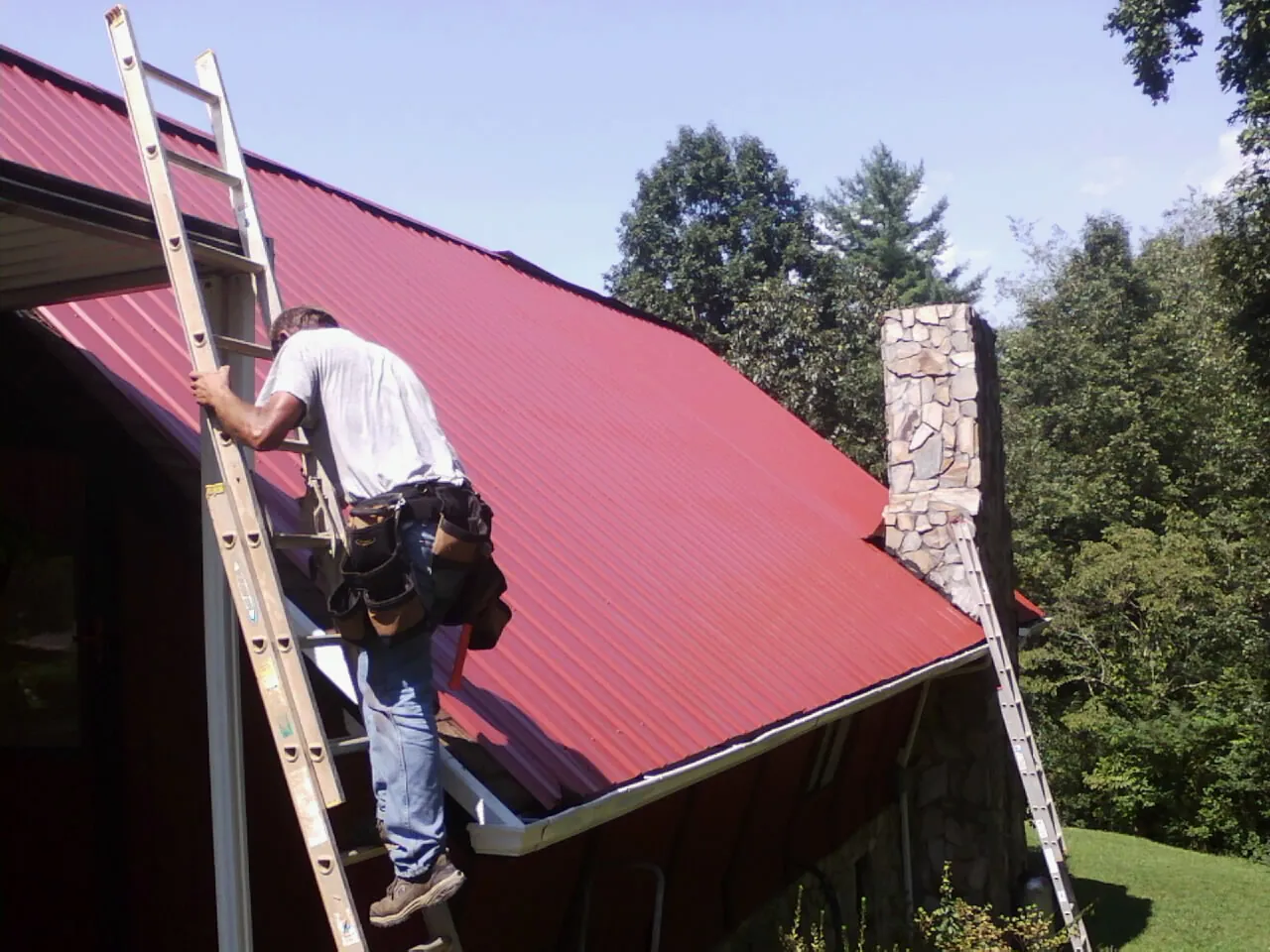 Roofer on a ladder beside a red metal roof and stone chimney — Soffit & Fascia Repair by Roofing Brothers in Rochester